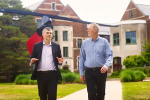 Two men walk and talk across a grassy campus courtyard in front of a brick academic building. One gestures with his hands while speaking, and a large red-and-black outdoor sculpture stands in the background among trees and landscaping.