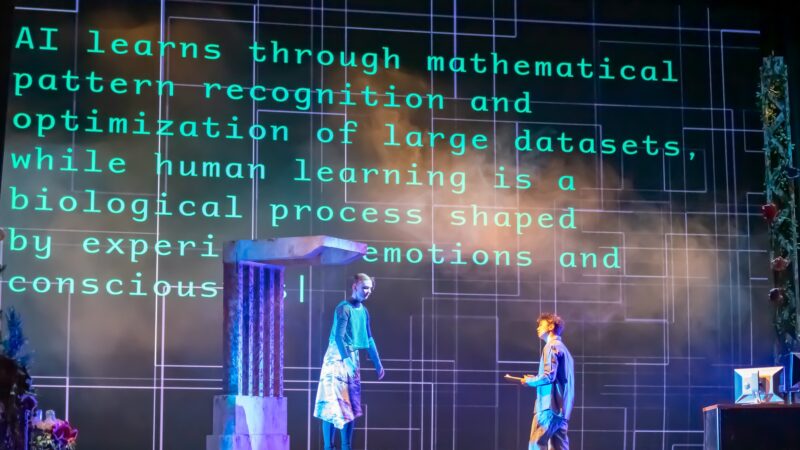 Two performers stand on a stage amid dramatic lighting while a large projection behind them displays text comparing artificial intelligence and human learning. The digital backdrop reads, "AI learns through mathematical pattern recognition and optimization of large datasets, while human learning is a biological process shaped by emotions and consciousness."
