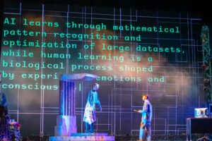 Two performers stand on a stage amid dramatic lighting while a large projection behind them displays text comparing artificial intelligence and human learning. The digital backdrop reads, "AI learns through mathematical pattern recognition and optimization of large datasets, while human learning is a biological process shaped by emotions and consciousness."