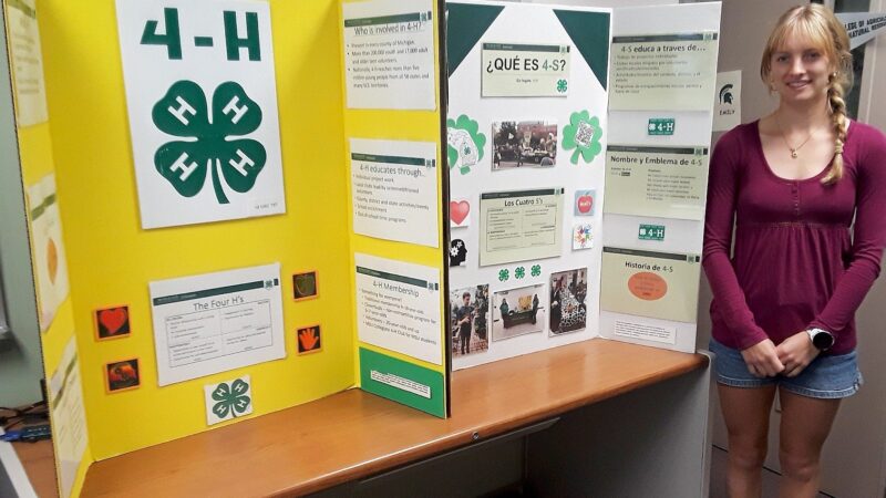 A student stands beside a tri-fold educational display about 4-H featuring bilingual English and Spanish information, photos, and the organization’s four-leaf clover emblem. The presentation board includes sections about 4-H history, membership, and programming, displayed on a table indoors.