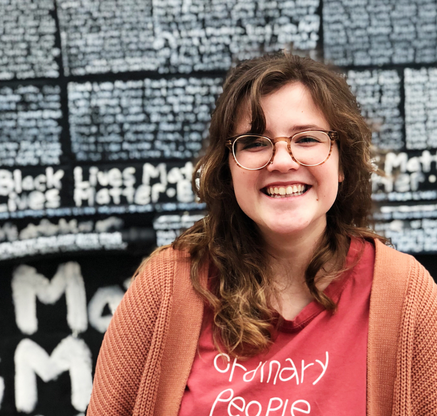 woman wearing a pink shirt smiling in front of a black wall with white text