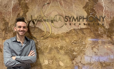 A man with blonde hair is standing with his arms crossed over his chest in front of a granite wall that reads, "The Virginia Symphony"