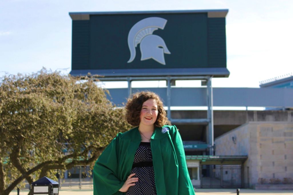 Woman wearing green graduation gown over a black dress standing in front of the spartan stadium
