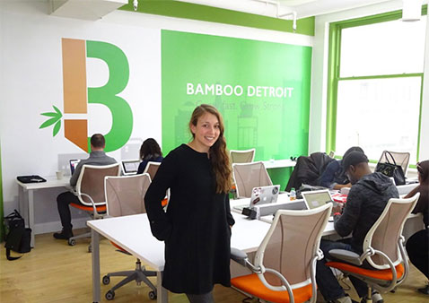 Woman with long brown hair wearing a black long sleeve dress standing in an office with a desk and chairs and a white, orange, and green wall that says "Bamboo Detroit" on it