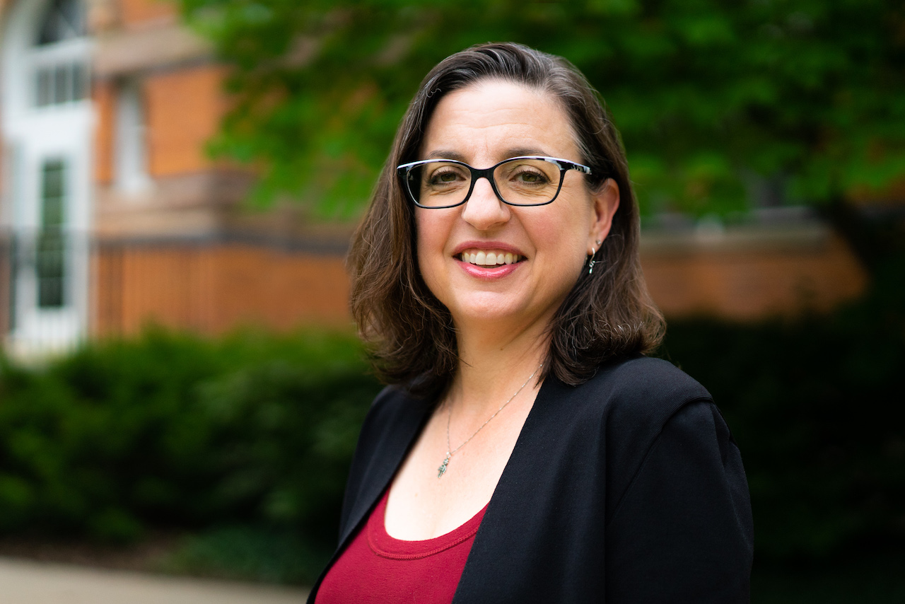 woman who is wearing a pink shirt, black blazer, and black glasses is smiling and standing in front of a brick building
