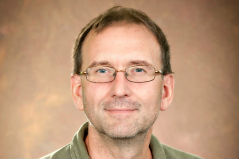 Man with short hair and glasses in a green polo against a brown background smiling at the camera