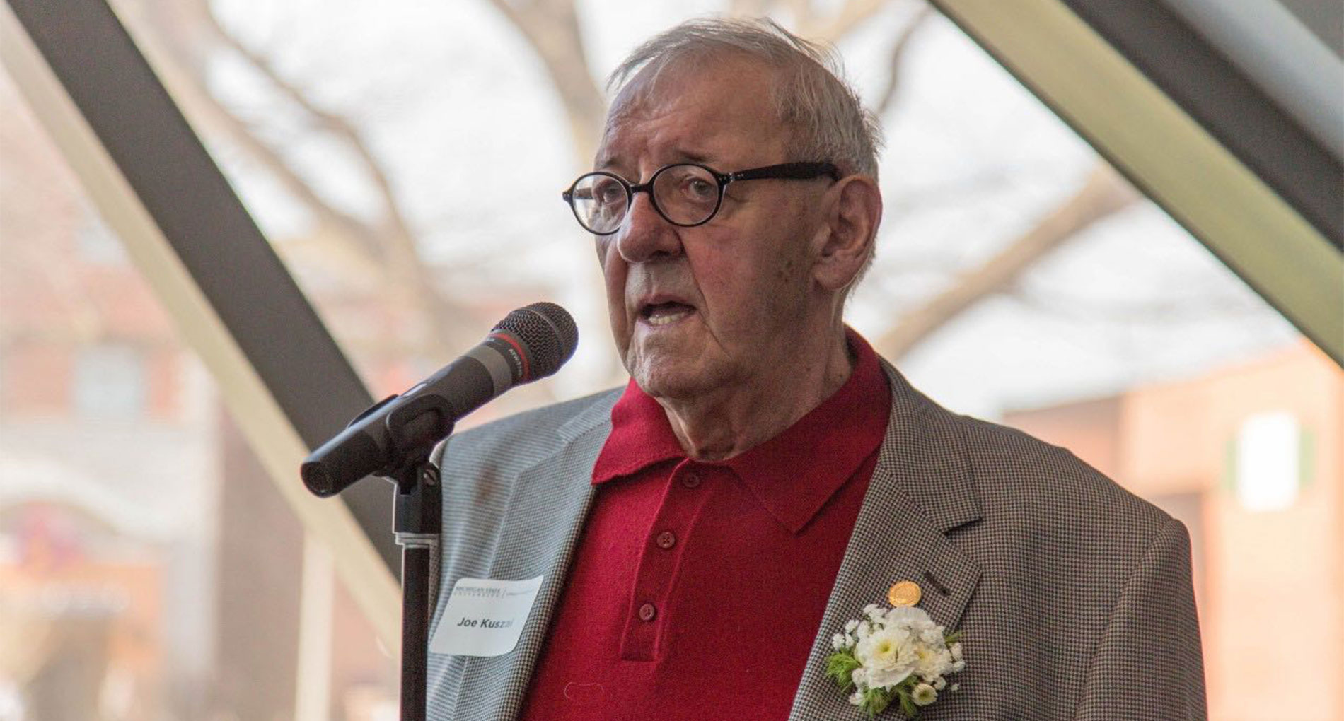 Man wearing checkered blazer with red shirt and glasses talking while standing in front of a microphone