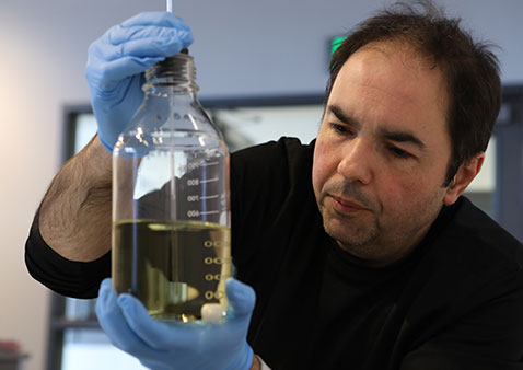Man with brown hair wearing a black shirt is examining a testing tube filled with yellow liquid. He is wearing blue rubber gloves