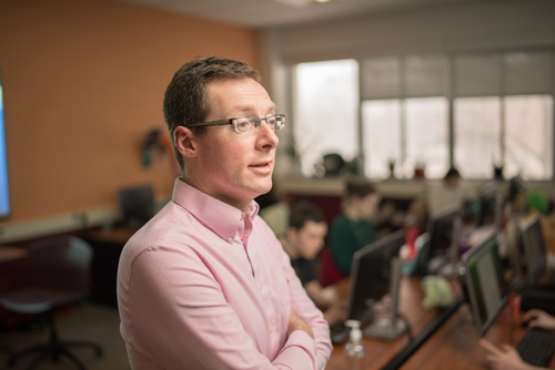 portrait of man with glasses in pink button up with classroom and students in background