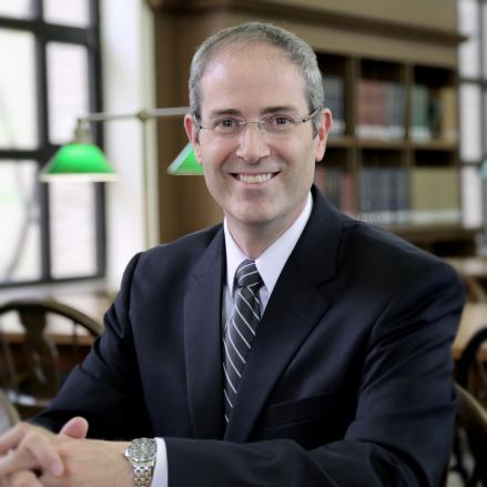 a man wearing glasses with a black suit, a white button down shirt and blue tie
