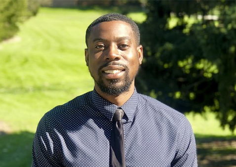 man wearing dark blue button-up with black tie who is smiling in front of trees