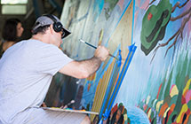 Man with headphones, a hat, and glasses painting a brightly colored mural.