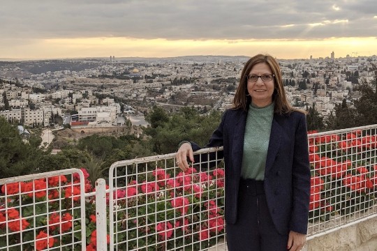 a photo of a woman with brown hair wearing glasses, a navy blue blazer, with a city behind her