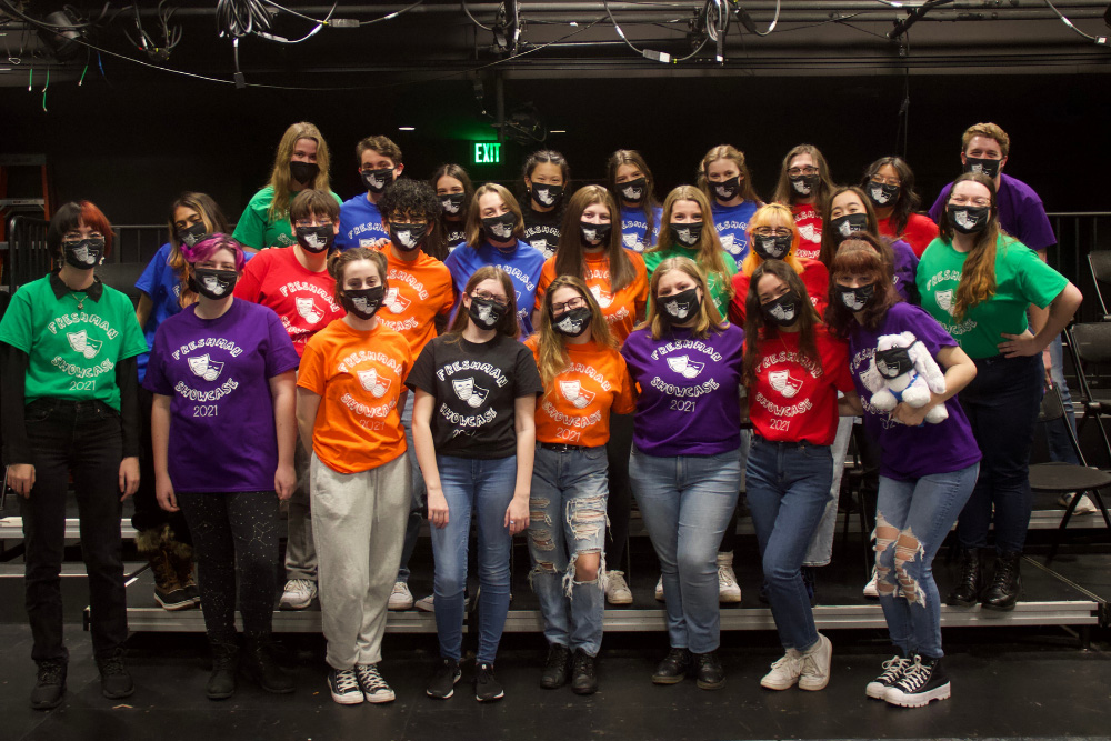 A group of students pose as a group in a theater. They wear different colored shirts, representing their different team.