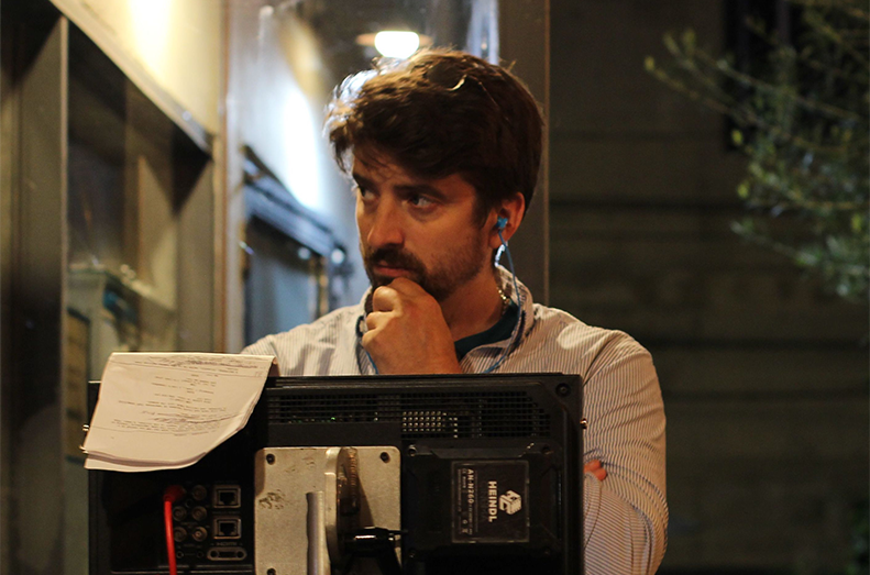 A man stands behind a screen with a notepad. His hand is near his chin and he has an ear bud in his ear.
