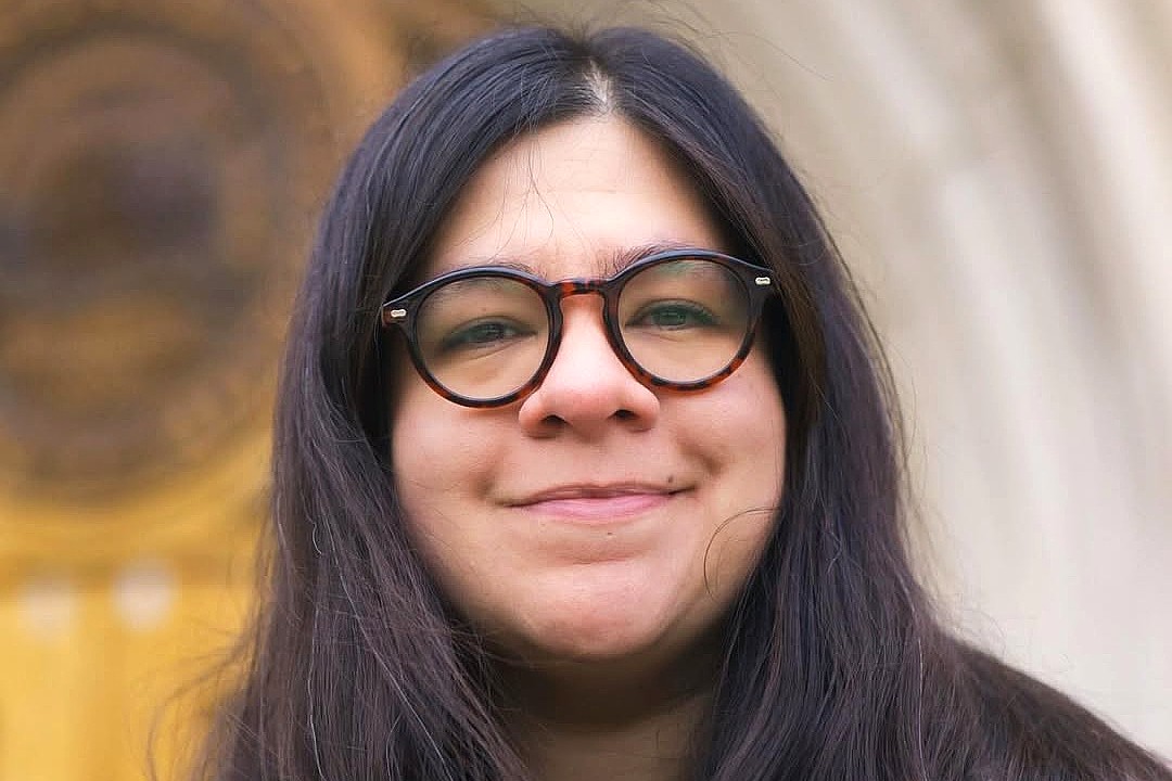 A close-up portrait of a woman with long dark hair and glasses, smiling gently. She is standing in front of a blurred background featuring an ornate architectural design with golden hues.