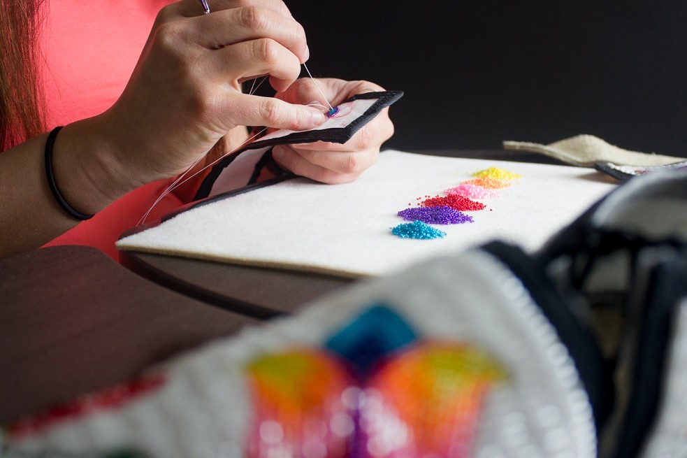 A close-up of a person beading by hand. Their hands are stitching black fabric while colorful piles of seed beads in a rainbow gradient sit on a white work surface.