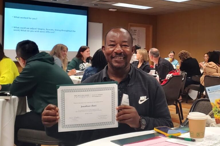 A man in a black jacket and black shirt sits at a table in a conference room, smiling and holding up a certificate from Michigan State University. He is surrounded by other attendees and a presentation slide is visible in the background.