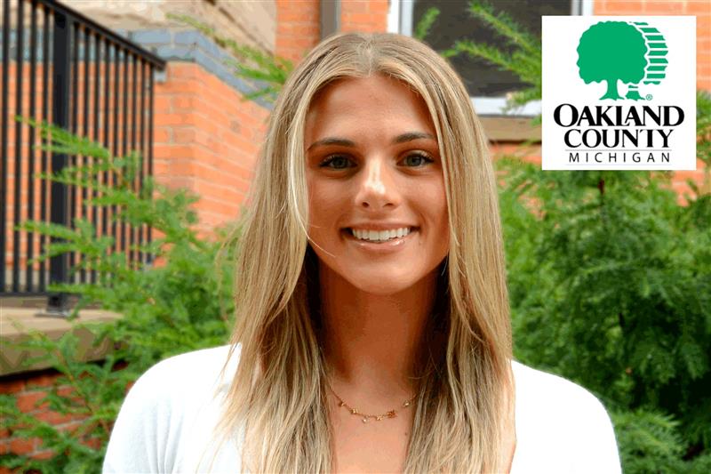 A person with long blonde hair and a white shirt smiling outdoors in front of greenery and a brick building, with the Oakland County, Michigan logo in the top right corner.