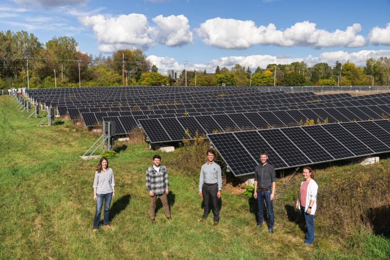 Five people stand in front of a large field of solar panels on a sunny day. Rows of panels stretch into the distance, surrounded by green grass and trees under a blue sky with scattered clouds