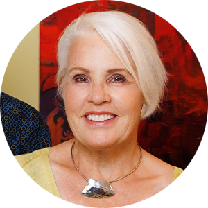 Woman with white hair smiles warmly, wearing a yellow top and statement necklace against a vibrant red and black abstract background.