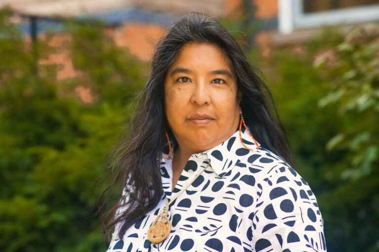 A woman with long dark hair stands outdoors in front of green foliage and a brick building. She wears a white collared shirt with a black geometric pattern and orange beaded earrings