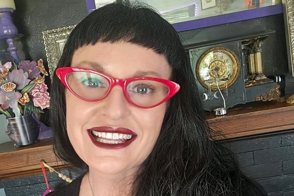 A person with long dark hair and bangs smiles at the camera, wearing bright red cat-eye glasses and dark lipstick. Behind them is a dark mantel decorated with flowers, framed art, and a vintage clock.