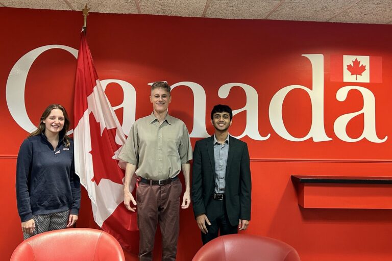 Three people stand side by side in front of a bright red wall featuring the word “Canada” in large white letters. A Canadian flag is between the person on the left and the person in the center. The person on the left wears a dark pullover and patterned pants, the person in the middle wears a short-sleeve button-down shirt and trousers, and the person on the right wears a dark suit with a light dress shirt. All three are smiling.