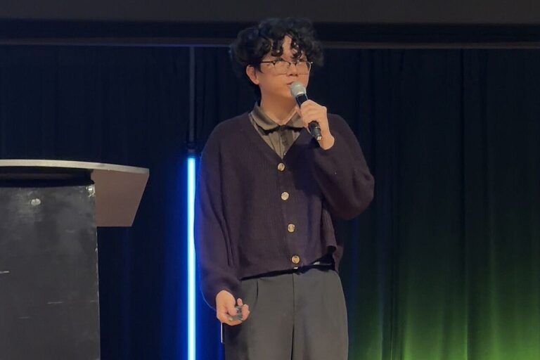 Man, dressed all in black with short black curly hair and glasses, holding and speaking into a microphone on a stage with a dark background.