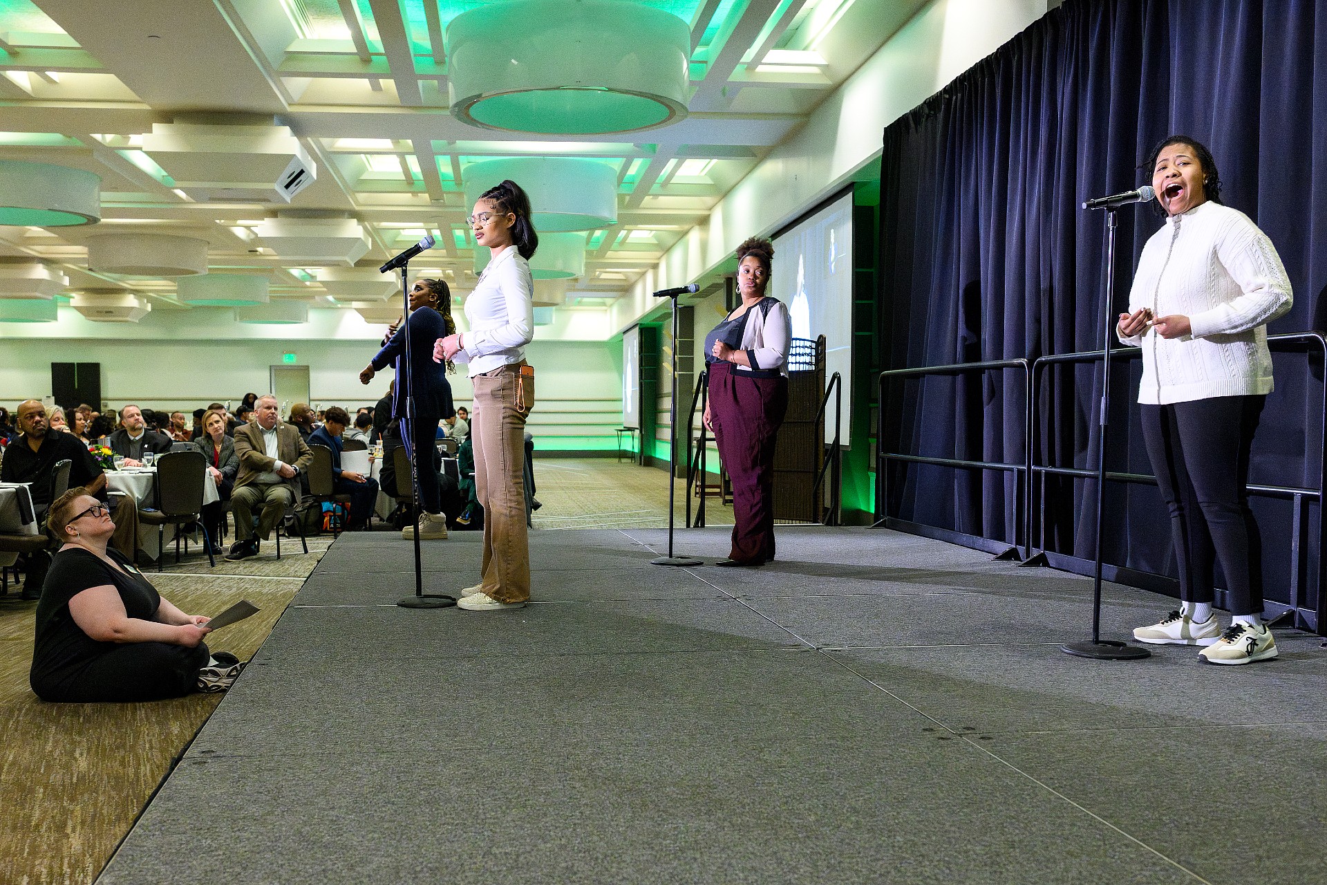 Tasiyah Hampton, Kamara Drane, and Charisma Holly stand on a raised stage, each at a microphone. They wear professional-casual attire in neutral tones. A dark curtain forms the stage backdrop, while audience members seated at round banquet tables fill the foreground in a large conference hall.