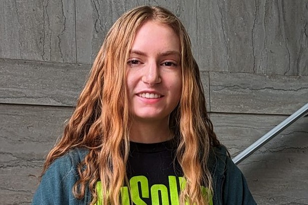 A professional headshot of Rachel Lewis. She has long, wavy light-brown hair and is wearing a dark teal zip-up hoodie over a black t-shirt featuring a lime-green "Museum" logo. She is smiling against a textured grey stone wall background.