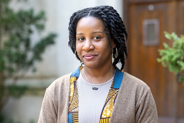 A professional headshot of Alexandria Davis, Assistant Professor of Dance at Michigan State University. Her hair is styled in twists, and she wears gold hoop earrings, a thin gold necklace with a dark pendant, and a tan cardigan over a ribbed grey top with a blue and yellow patterned trim. She is looking toward the camera with a neutral, poised expression.