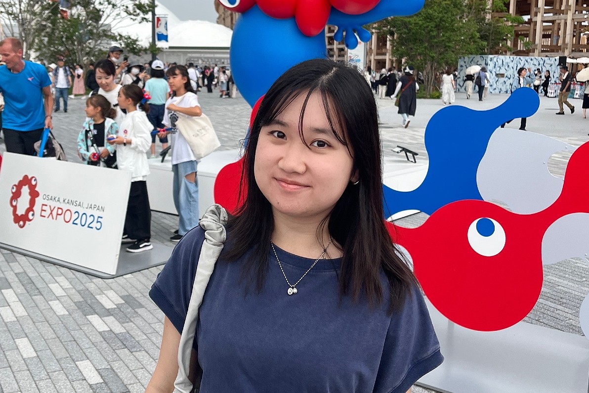 A young woman stands outdoors at a busy plaza, smiling slightly at the camera, with colorful red-and-blue Expo 2025 signage and a large cartoon-style mascot sculpture behind her as visitors walk through the space.