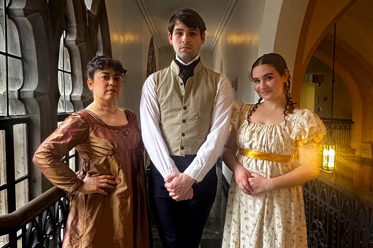 Three people in historical costumes stand in a corridor with arched windows. The mood is formal and posed, with warm lighting highlighting their attire.
