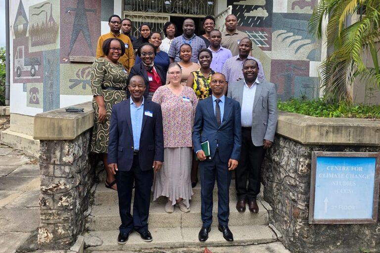 Workshop facilitators and participants pose together on the steps of a building bearing a sign for the Centre for Climate Change Studies at the University of Dar es Salaam, framed by a colorful painted mural and tropical greenery.