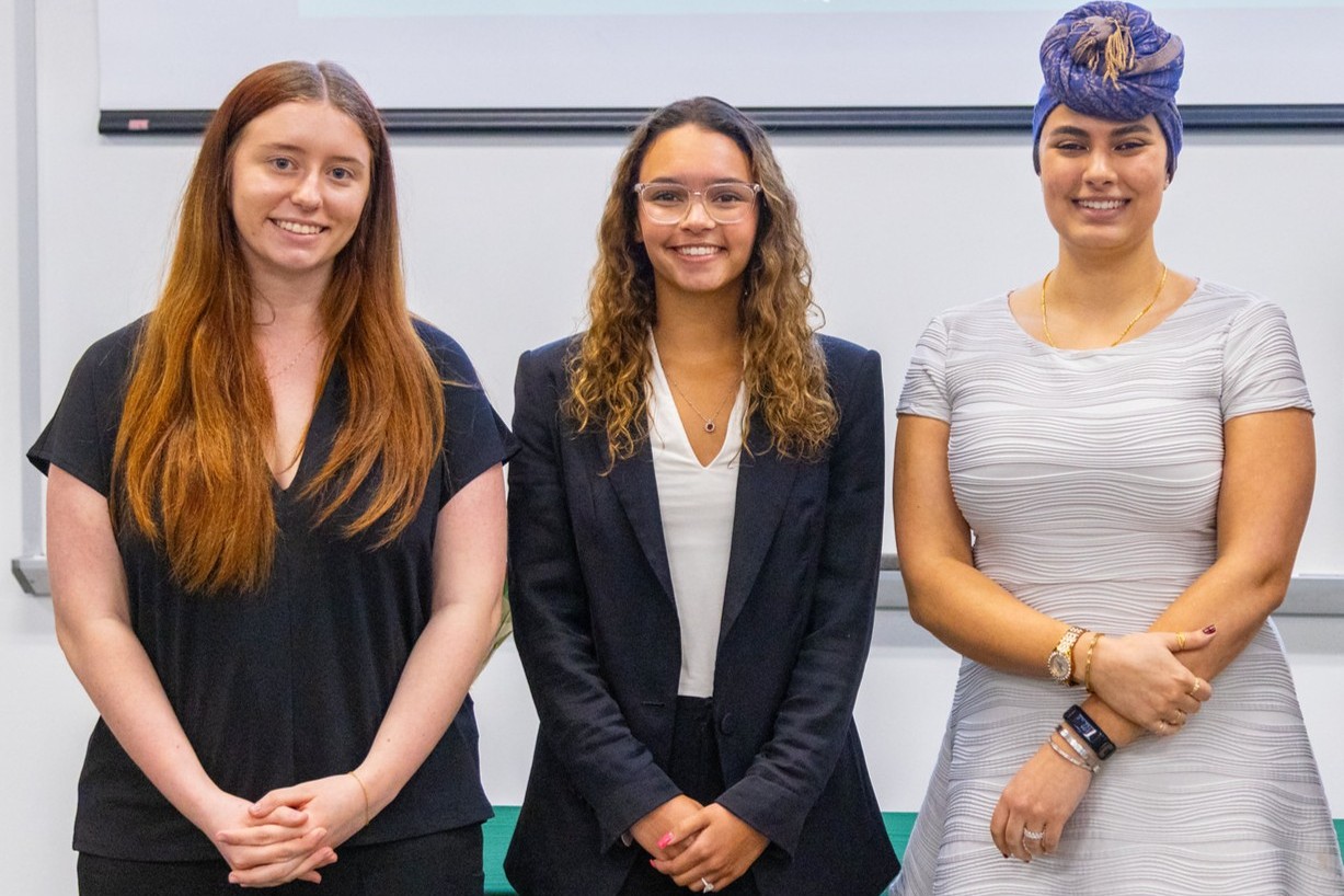 2026 award recipients Avery Riehl (left), Emma Wilkins (center), and Alliah Woodside (right) stand together smiling in front of a projected College of Arts & Letters slide. Riehl wears a black short-sleeve top with long red hair down; Wilkins wears a black blazer over a white top with curly hair and clear-framed glasses; Woodside wears a white ribbed fitted dress and an ornate blue and gold head wrap. A green Department of Religious Studies table is partially visible in the foreground.