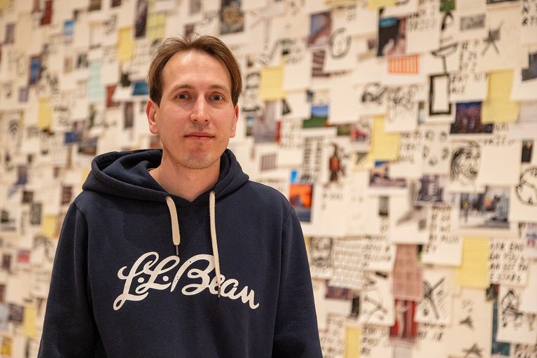 A man in a blue L.L. Bean hoodie stands in front of a wall filled with assorted notes and photos, conveying a creative and thoughtful atmosphere.