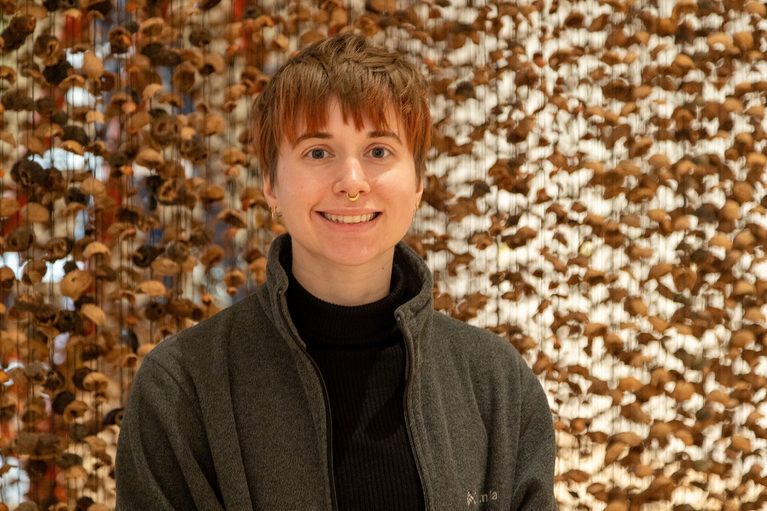 A person with short hair and a septum ring smiles warmly, wearing a dark jacket. Behind them, a textured background of dried leaves creates an earthy atmosphere.