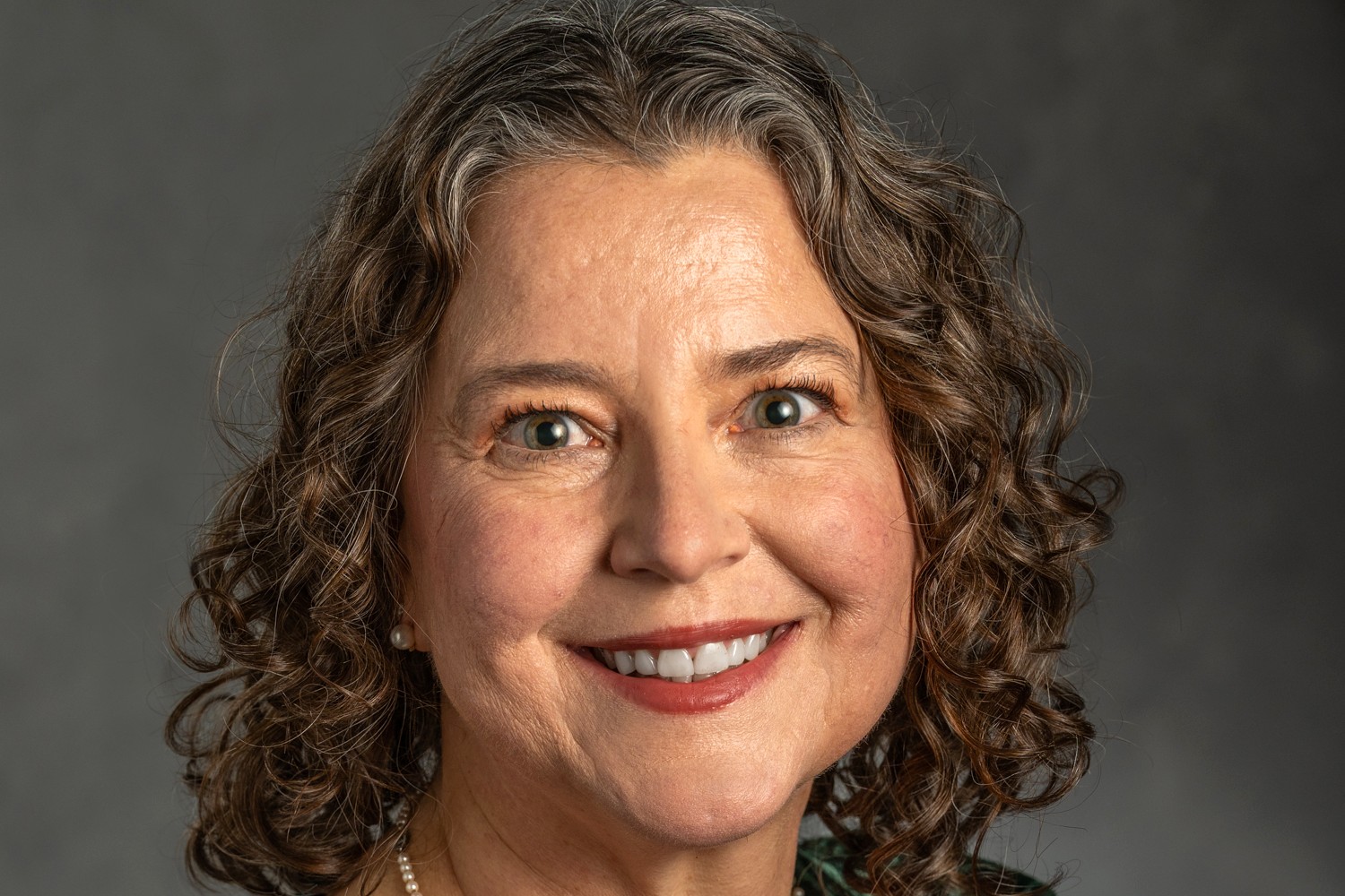 Smiling woman with curly hair in front of a gray background. Her expression is warm and friendly, wearing a subtle pearl earring and necklace.