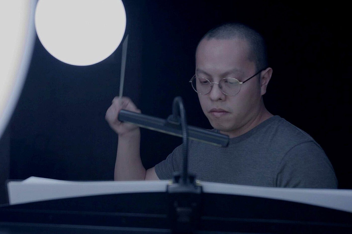 Austin Oting Har sits in a darkened recording studio, focused on a music stand holding sheet music while holding a percussion mallet. A bright studio light illuminates the scene as he prepares to perform or record music.