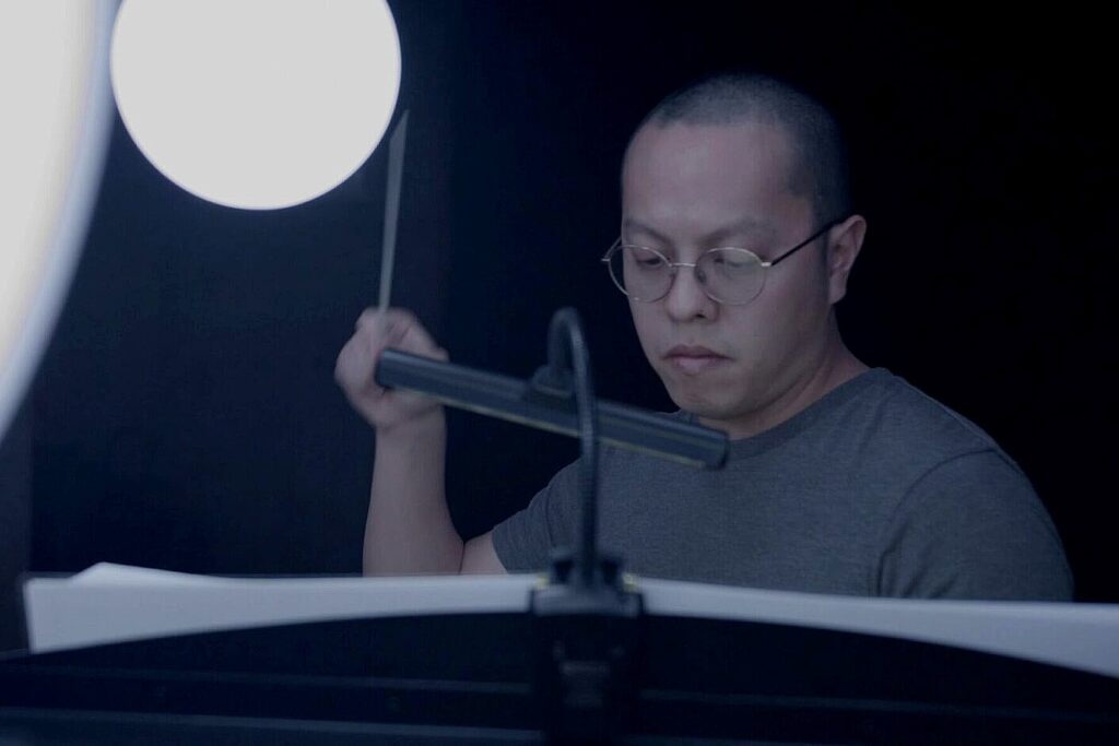 Austin Oting Har sits in a darkened recording studio, focused on a music stand holding sheet music while holding a percussion mallet. A bright studio light illuminates the scene as he prepares to perform or record music.