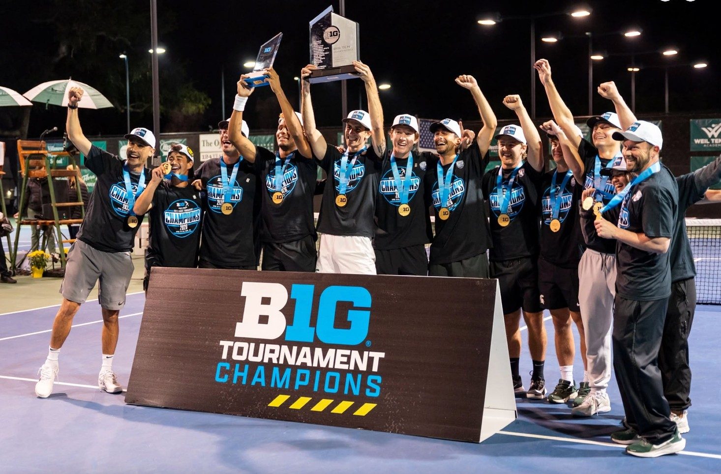 Michigan State men's tennis players and coaches celebrate on the court after winning the Big Ten Tournament championship, wearing championship T-shirts, medals, and hats while raising the championship trophy behind a "B1G Tournament Champions" sign.