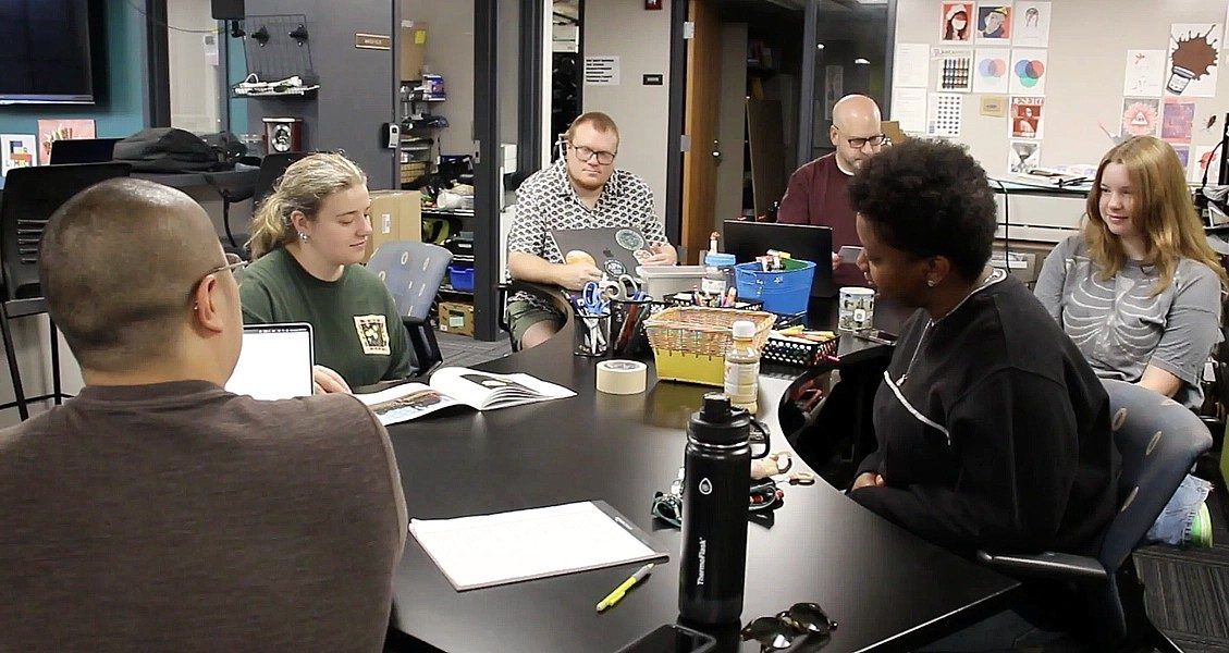 Austin Oting Har meets with a group of students around a conference table in the Bogue Street Records workspace. Laptops, notebooks, and design materials are spread across the table as the team collaborates on projects for the student-run record label.