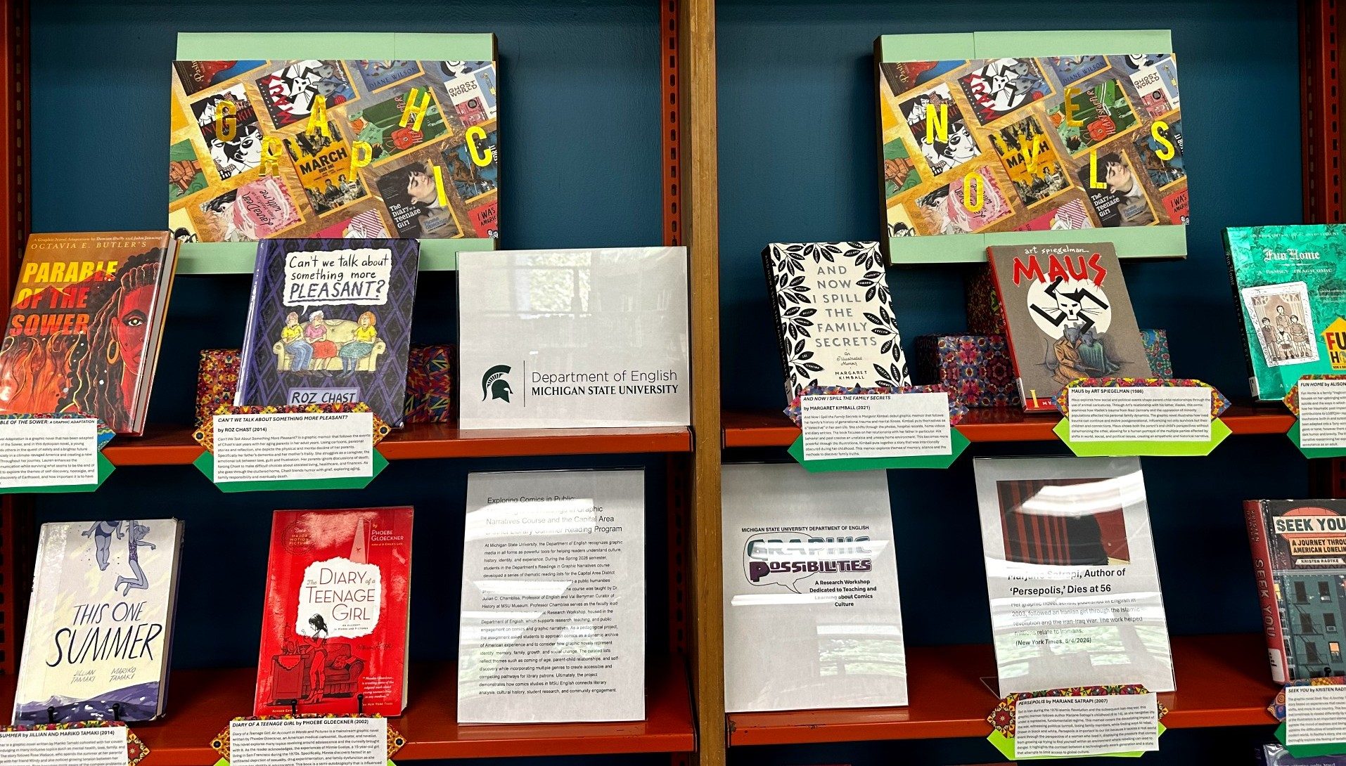 A display of graphic novels and comics-themed books arranged on orange shelving highlights a collaborative reading exhibit by Michigan State University’s Department of English. Featured titles include Parable of the Sower, Can’t We Talk About Something More Pleasant?, Maus, This One Summer, Diary of a Teenage Girl, and Seek You, accompanied by descriptive placards and informational signage about graphic narratives and community reading. Decorative boards spelling “GRAPHIC” and “NOVELS” frame the display above.