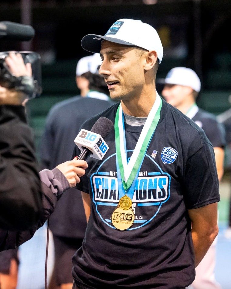 Michigan State men's tennis coach Harry Jadun, wearing a Big Ten Tournament champions T-shirt, championship medal, and white championship hat, speaks with a Big Ten Network reporter during an on-court post-match interview. Teammates celebrate in the background.