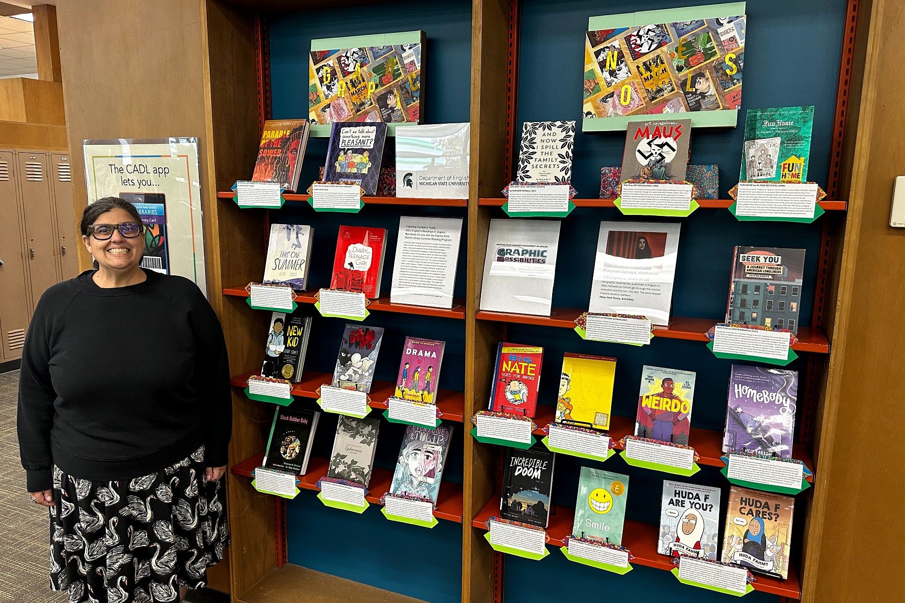 A smiling person stands beside a library display of colorful graphic novels arranged on shelves. Each book has a green label, creating a welcoming atmosphere.