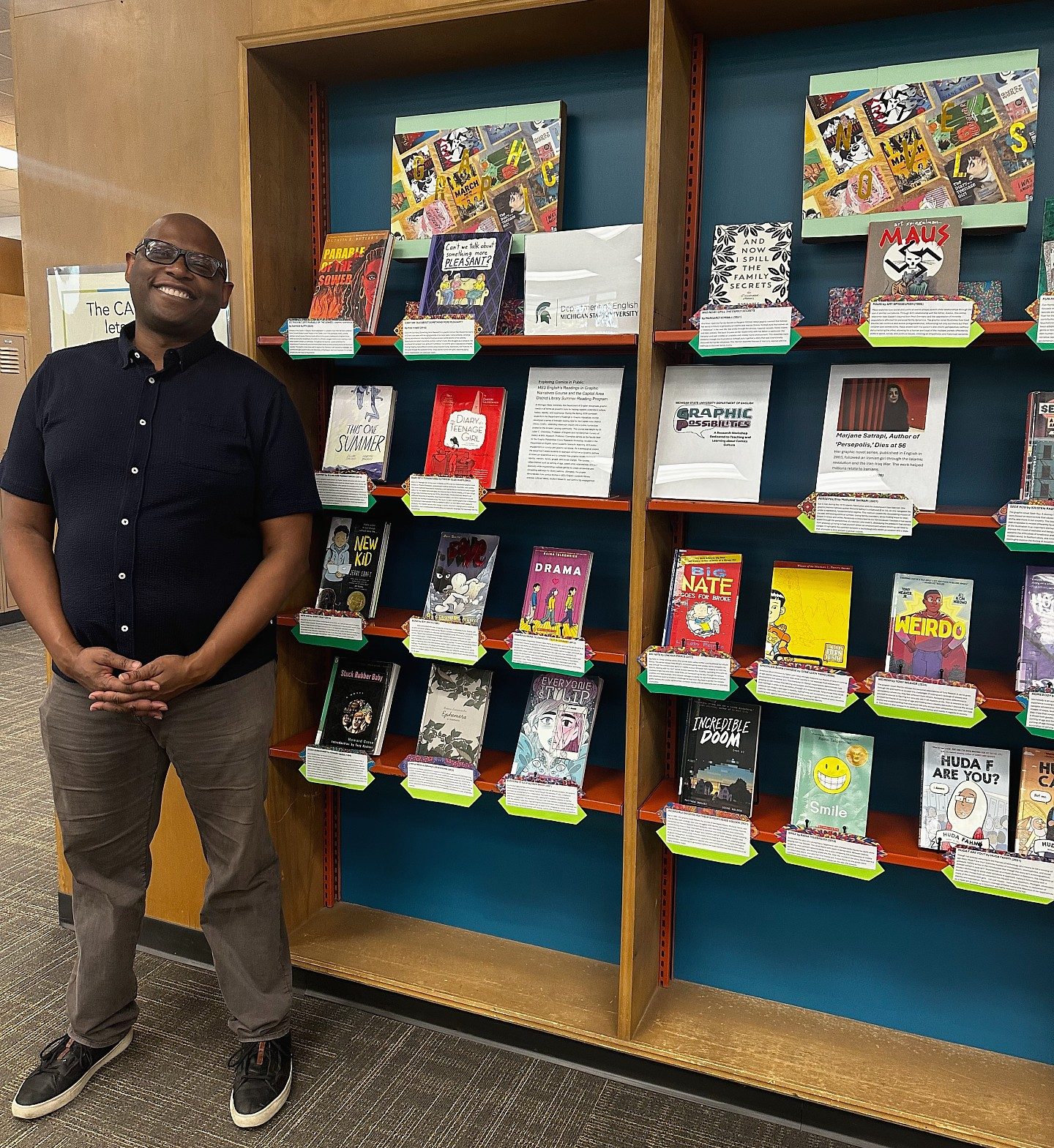 A person smiling stands beside a library book display featuring various graphic novels. The shelves are filled with colorful covers, creating a vibrant and welcoming atmosphere.