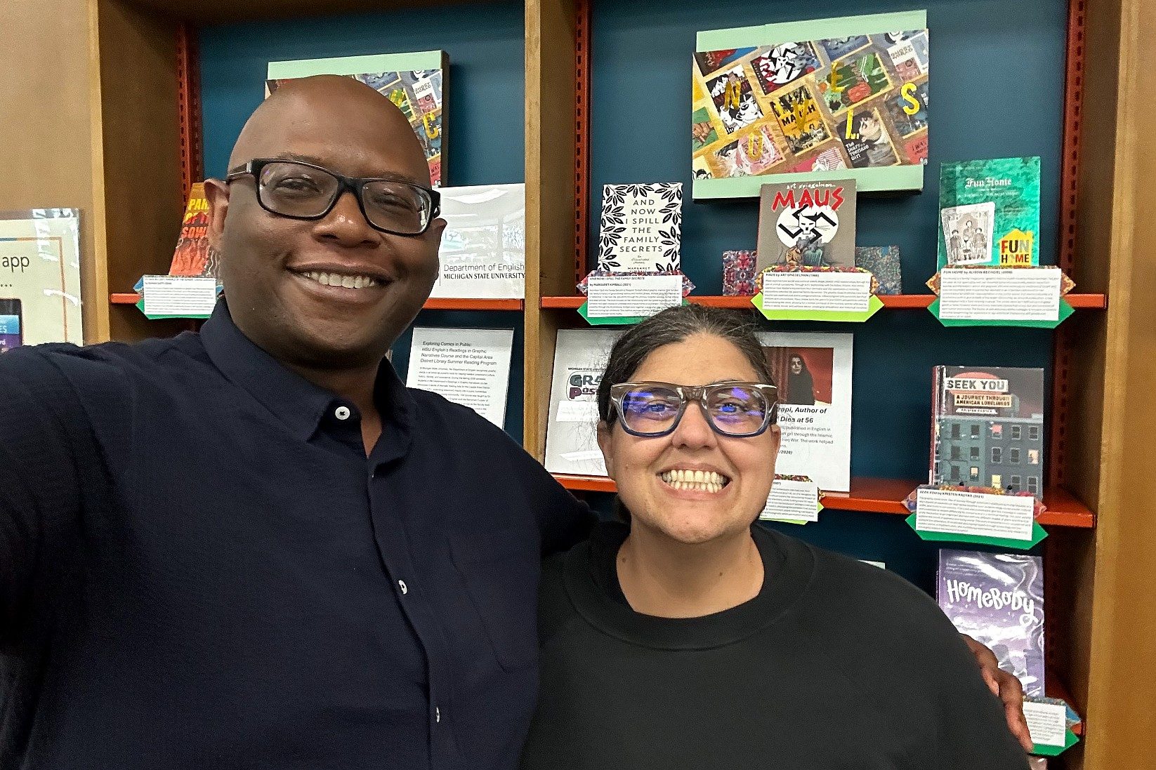 Two smiling people stand arm in arm in front of a library-style display featuring graphic novels and informational cards. Behind them, books including Maus, Fun Home, Seek You, and Homebody are arranged on shelves as part of a comics-themed exhibit.