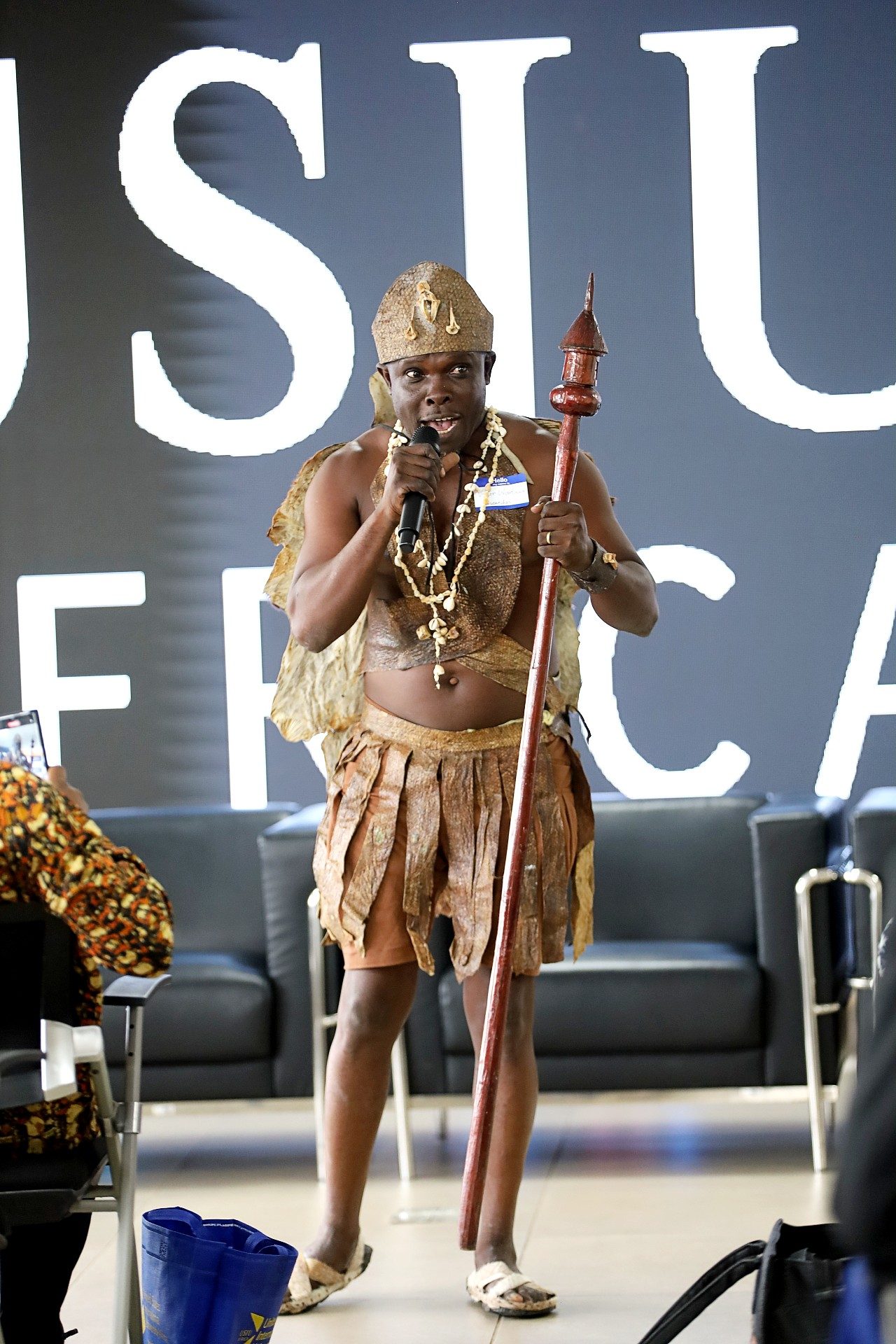 A workshop participant wearing traditional attire and ceremonial beads speaks into a microphone while holding a carved wooden staff during the “Documentation and Scholarly Publishing of Kenyan Indigenous Languages” workshop at United States International University-Africa in Nairobi, Kenya. Audience members are seated in the foreground as the participant delivers a cultural presentation.