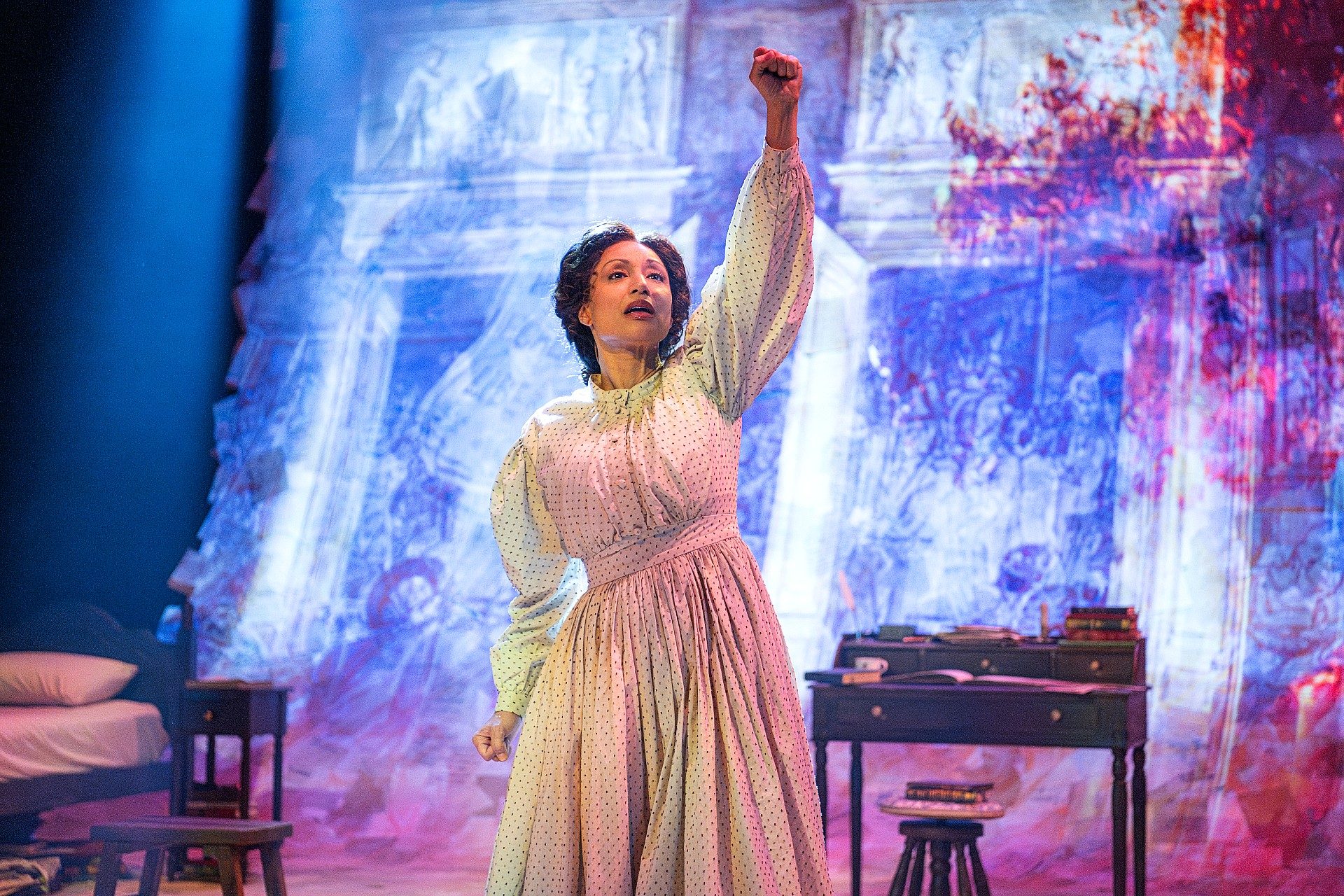 An actor in a long white period dress performs on stage, raising a clenched fist overhead while standing in front of a colorful projected backdrop. A bed, desk, books, and other furnishings surround the performer, creating the setting of a historical drama. Dramatic lighting highlights the actor’s expressive pose and determined expression.